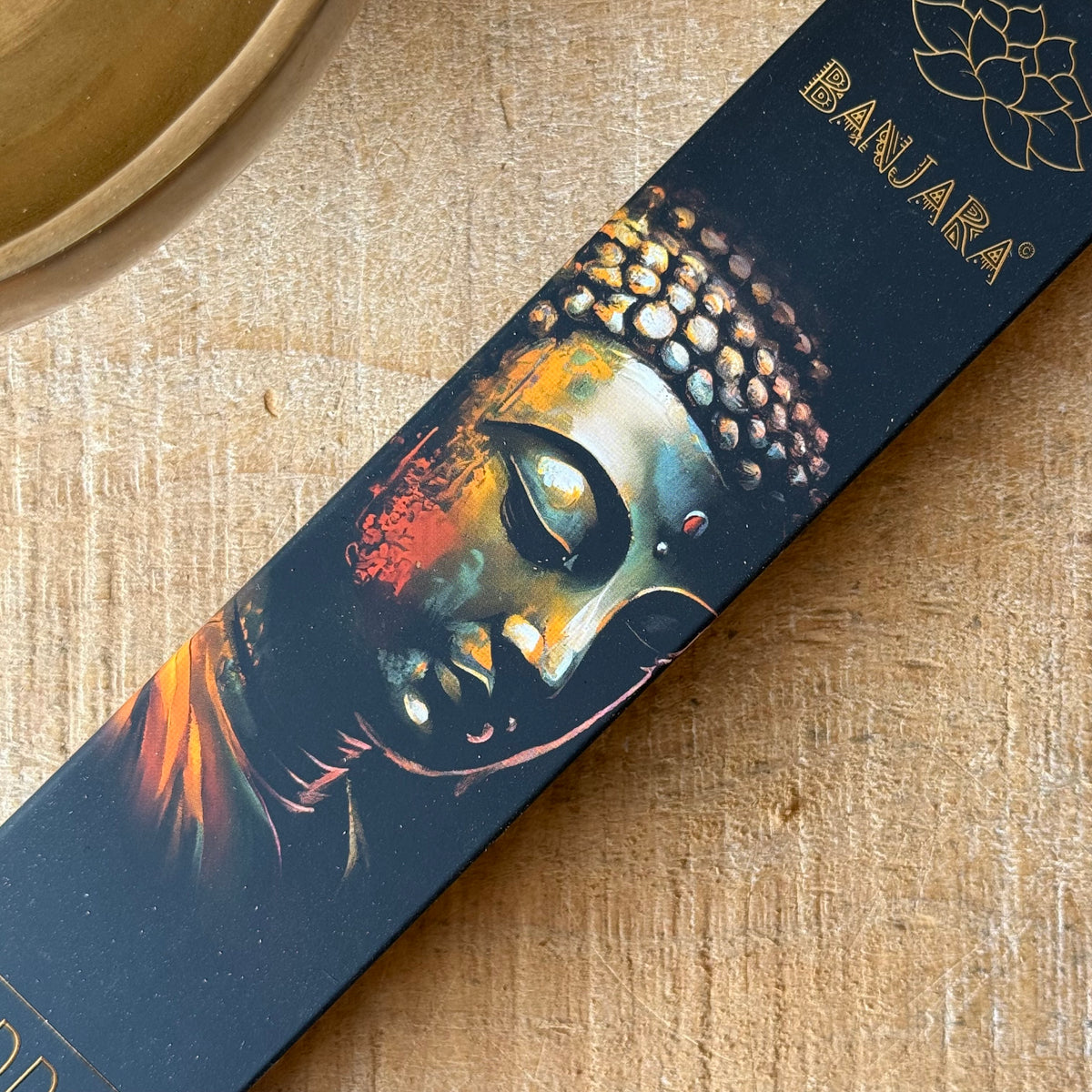 A close-up of a rectangular product box featuring a stylized Buddha face illustration in warm orange, gold, and teal tones on a dark background. The brand name 'BANJARA' appears in gold near the top right corner.