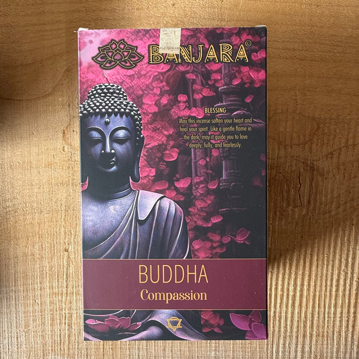 A rectangular product box for Banjara Buddha Compassion incense sticks, featuring a dark blue Buddha statue and pink lotus petals on a wooden surface.