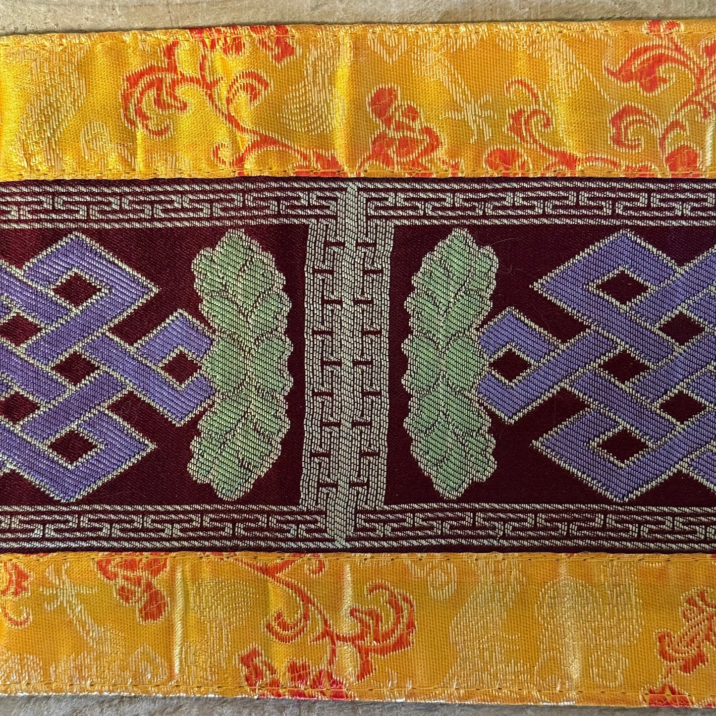 This Tibetan Ritual Mat features the sacred knot of infinity, a Buddhist symbol representing the interdependence of all phenomena and the development of wisdom. Perfectly designed for altars, singing bowls, or placing sacred ornaments, this high-quality altar cloth is made from thick silk brocade and features an embroidered dorje at its center, surrounded by a self-lotus design. Measuring 35 x 15 cm, it adds both elegance and spiritual significance to your meditation or ritual space. Handcrafted in Nepal, e