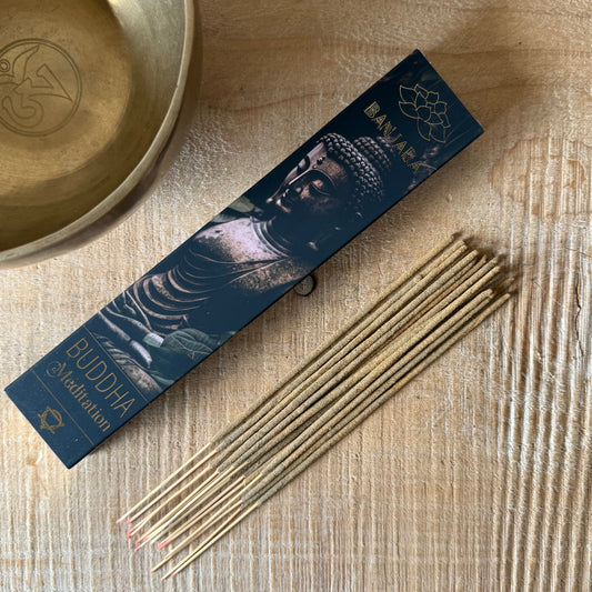 A box of Baniar Buddha Meditation incense sticks lies on a light wood surface next to several incense sticks, with a brass bowl partially visible in the top left corner.
