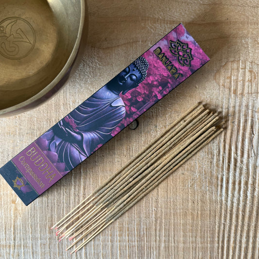Buddha incense sticks in a purple and pink box labeled 'Buddha Compassion' and 'Mantra', arranged on a light wood surface next to a brass bowl.