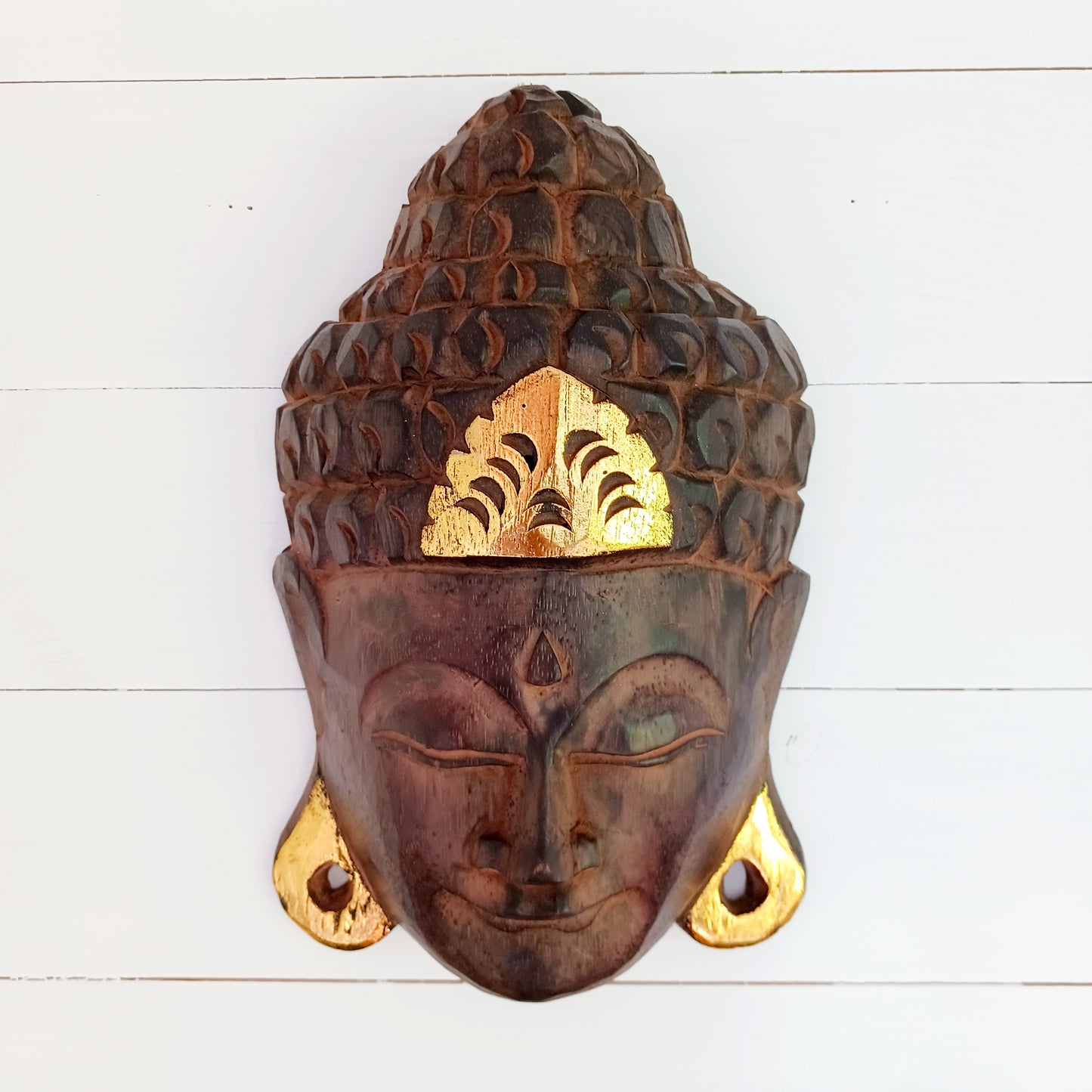 This skillfully crafted wall art of Lord Buddha with its beautiful, serene expression is both rustic and well-polished,  This mask has been hand carved from rain-tree wood by Indonesian artisans and is finished with gold leaf effect embellishment.&nbsp;  As this item is hand carved slight variations should be expected.  Size: Approx: 19cm × 28cm × 9cm Handmade and Fairtrade from Indonesia