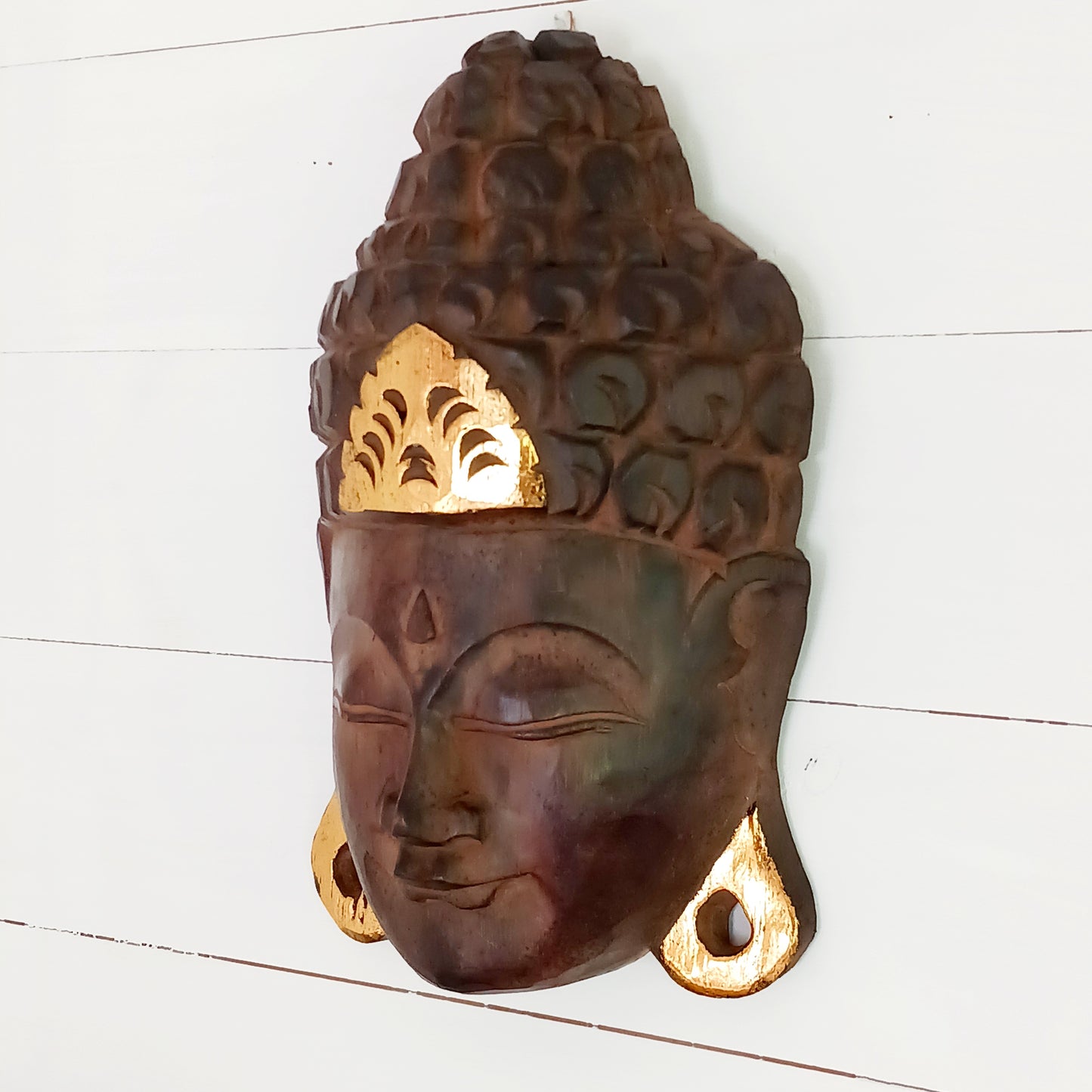 This skillfully crafted wall art of Lord Buddha with its beautiful, serene expression is both rustic and well-polished,  This mask has been hand carved from rain-tree wood by Indonesian artisans and is finished with gold leaf effect embellishment.&nbsp;  As this item is hand carved slight variations should be expected.  Size: Approx: 19cm × 28cm × 9cm Handmade and Fairtrade from Indonesia