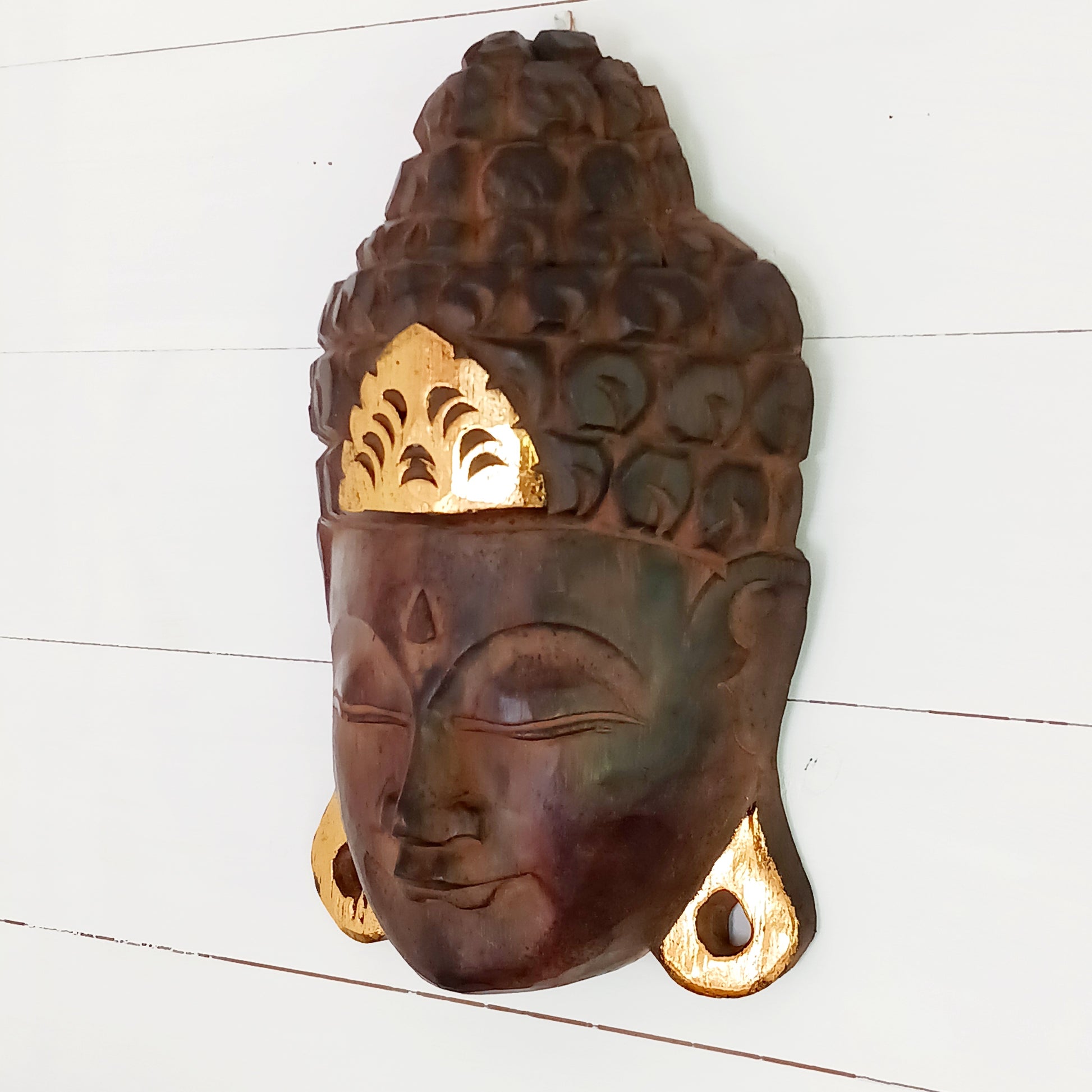 This skillfully crafted wall art of Lord Buddha with its beautiful, serene expression is both rustic and well-polished,  This mask has been hand carved from rain-tree wood by Indonesian artisans and is finished with gold leaf effect embellishment.&nbsp;  As this item is hand carved slight variations should be expected.  Size: Approx: 19cm × 28cm × 9cm Handmade and Fairtrade from Indonesia