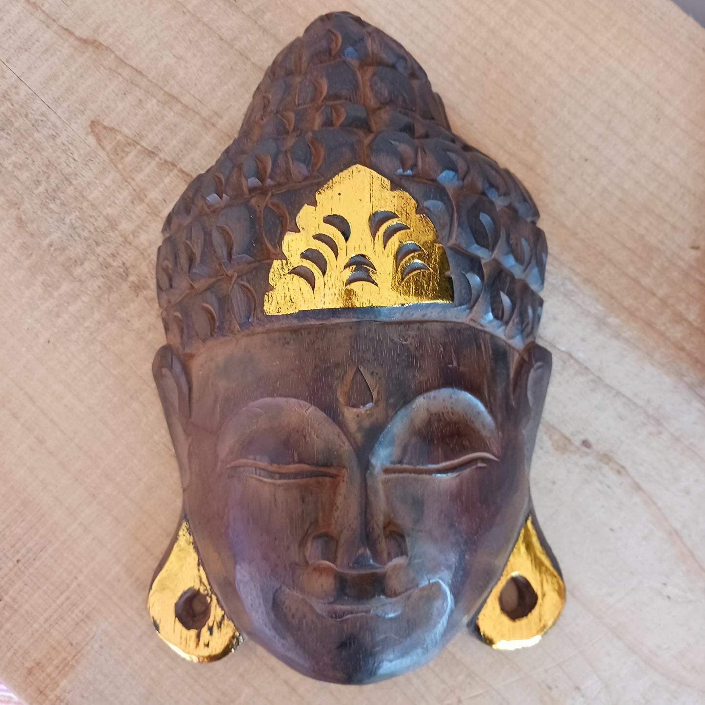 This skillfully crafted wall art of Lord Buddha with its beautiful, serene expression is both rustic and well-polished,  This mask has been hand carved from rain-tree wood by Indonesian artisans and is finished with gold leaf effect embellishment.&nbsp;  As this item is hand carved slight variations should be expected.  Size: Approx: 19cm × 28cm × 9cm Handmade and Fairtrade from Indonesia