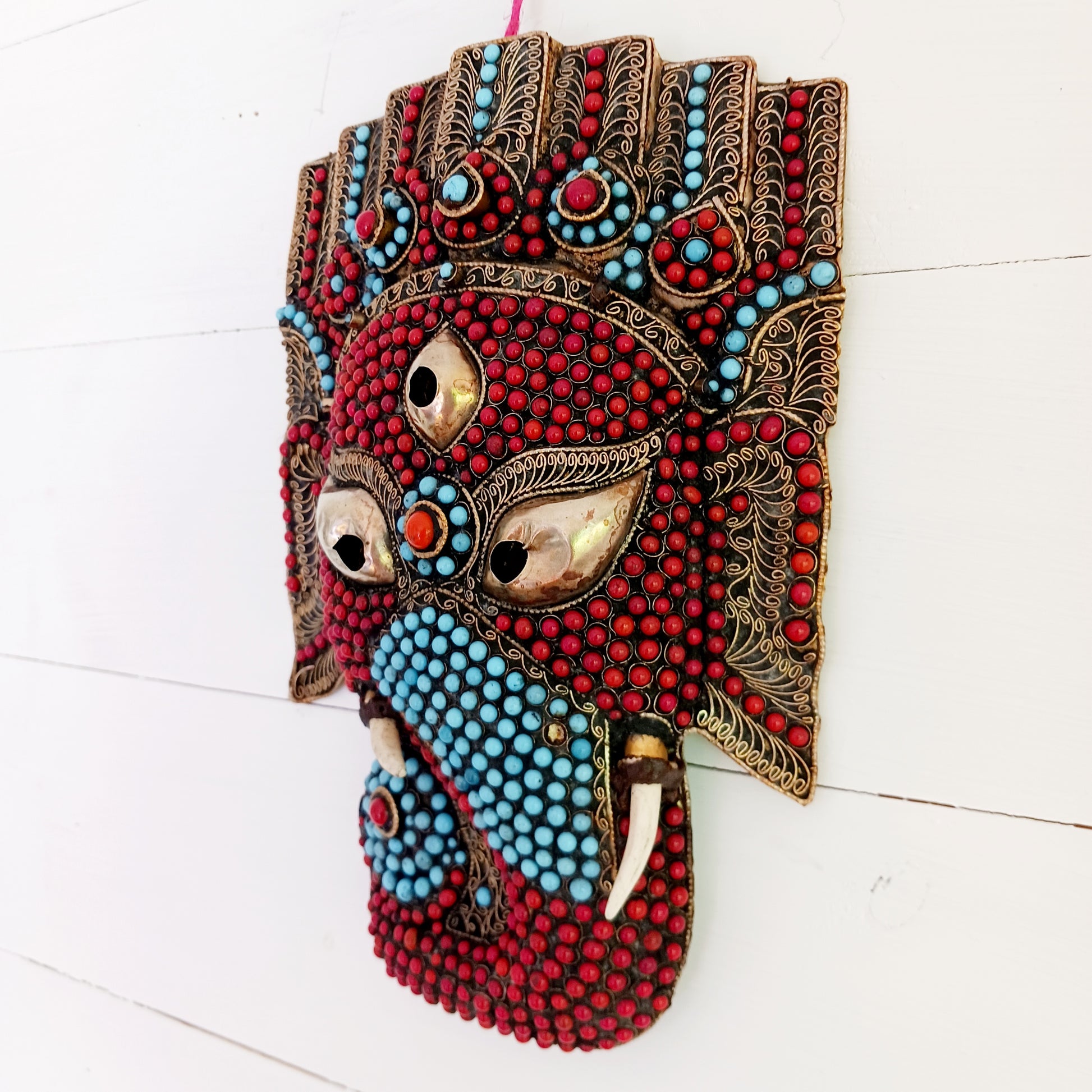 This stunning vintage handmade Lord Ganesh wearable mask | wall decor has multi stone embellishment detail. The colourful mask has hundreds of coral and turquoise stones set into brass wire-scrolls in the cloisonne manner. The third eye of insight is central to the mask. The piece is finished with beautiful, handcrafted filigree.