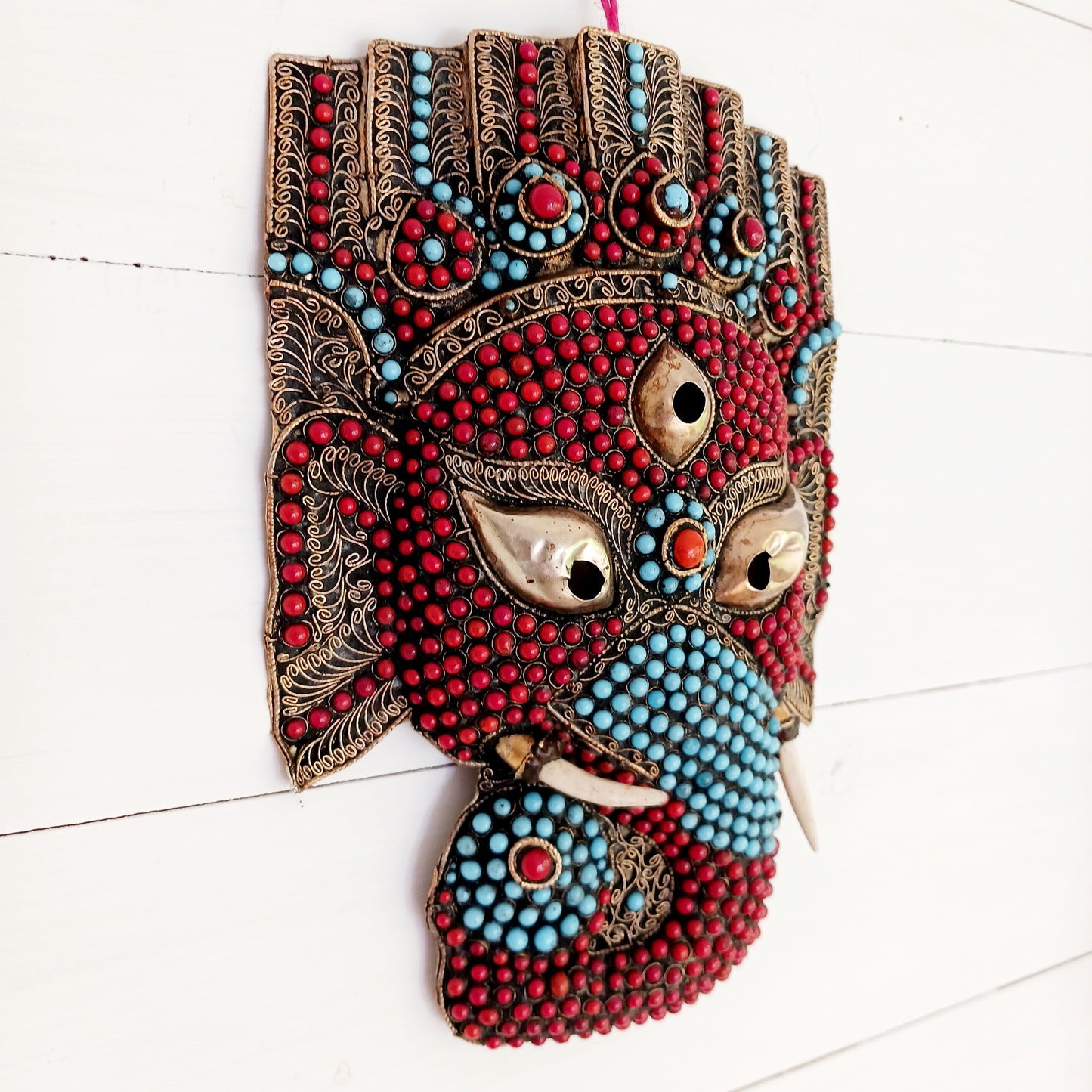 This stunning vintage handmade Lord Ganesh wearable mask | wall decor has multi stone embellishment detail. The colourful mask has hundreds of coral and turquoise stones set into brass wire-scrolls in the cloisonne manner. The third eye of insight is central to the mask. The piece is finished with beautiful, handcrafted filigree.