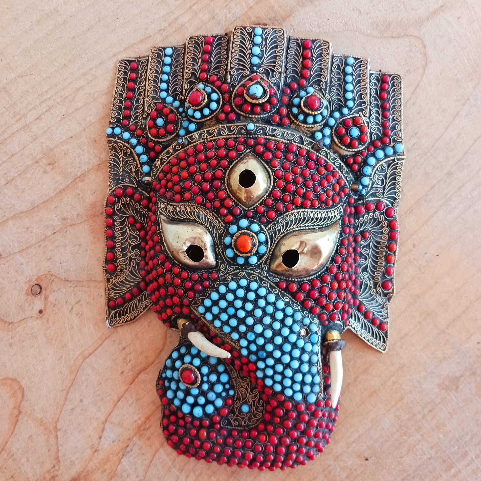 This stunning vintage handmade Lord Ganesh wearable mask | wall decor has multi stone embellishment detail. The colourful mask has hundreds of coral and turquoise stones set into brass wire-scrolls in the cloisonne manner. The third eye of insight is central to the mask. The piece is finished with beautiful, handcrafted filigree.