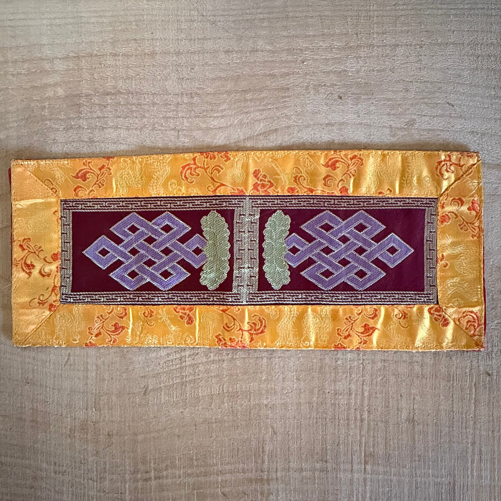 This Tibetan Ritual Mat features the sacred knot of infinity, a Buddhist symbol representing the interdependence of all phenomena and the development of wisdom. Perfectly designed for altars, singing bowls, or placing sacred ornaments, this high-quality altar cloth is made from thick silk brocade and features an embroidered dorje at its center, surrounded by a self-lotus design. Measuring 35 x 15 cm, it adds both elegance and spiritual significance to your meditation or ritual space. Handcrafted in Nepal, e