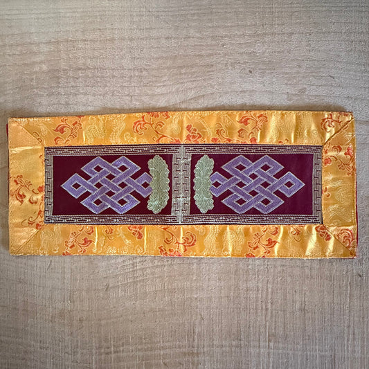 This Tibetan Ritual Mat features the sacred knot of infinity, a Buddhist symbol representing the interdependence of all phenomena and the development of wisdom. Perfectly designed for altars, singing bowls, or placing sacred ornaments, this high-quality altar cloth is made from thick silk brocade and features an embroidered dorje at its center, surrounded by a self-lotus design. Measuring 35 x 15 cm, it adds both elegance and spiritual significance to your meditation or ritual space. Handcrafted in Nepal, e