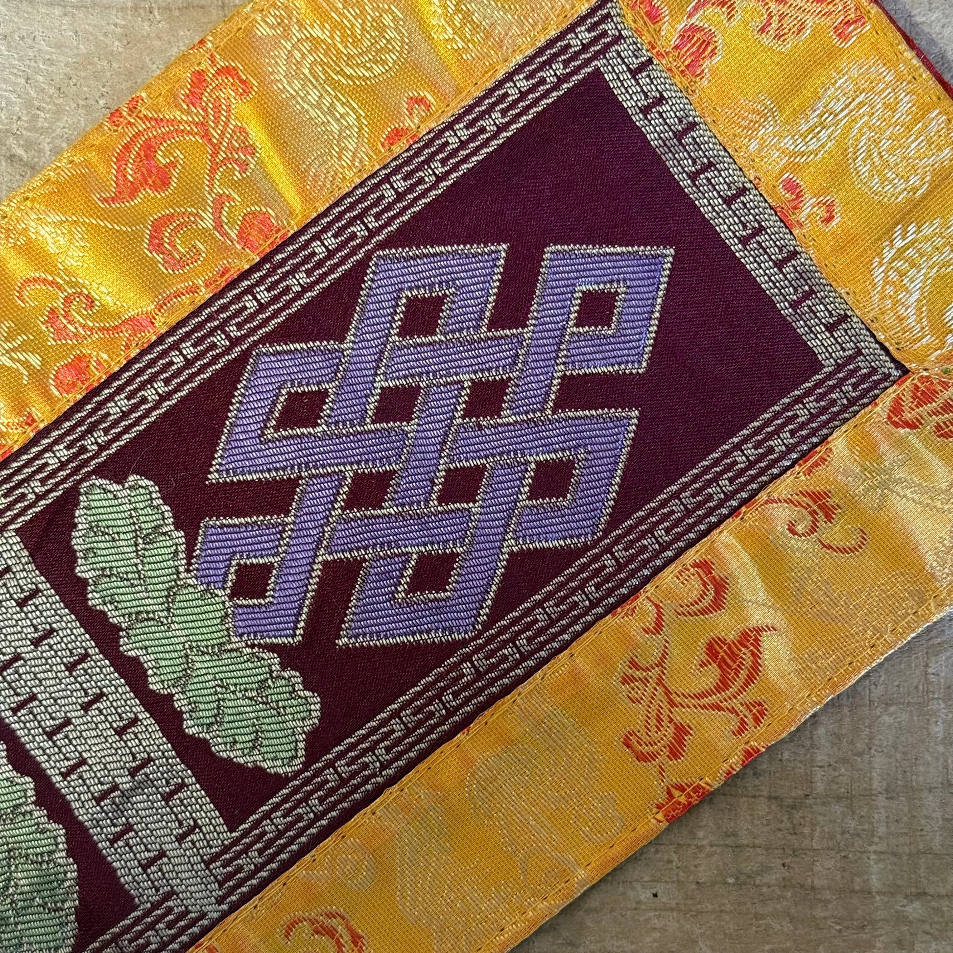 This Tibetan Ritual Mat features the sacred knot of infinity, a Buddhist symbol representing the interdependence of all phenomena and the development of wisdom. Perfectly designed for altars, singing bowls, or placing sacred ornaments, this high-quality altar cloth is made from thick silk brocade and features an embroidered dorje at its center, surrounded by a self-lotus design. Measuring 35 x 15 cm, it adds both elegance and spiritual significance to your meditation or ritual space. Handcrafted in Nepal, e