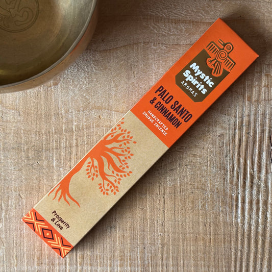 A box of Mystic Spirits Aromas Palo Santo & Cinnamon incense sticks lies diagonally on a light wood surface, with a brass bowl partially visible in the top-left corner.