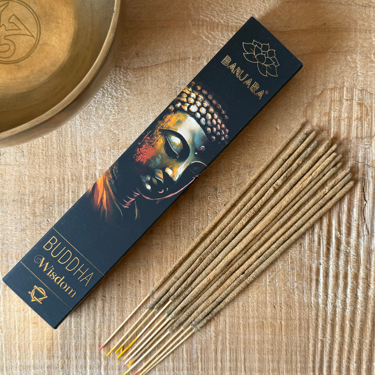 A box of Banjara Buddha Wisdom incense sticks lies diagonally on a light wood surface next to several unbaked incense sticks. A wooden bowl is partially visible in the top-left corner.