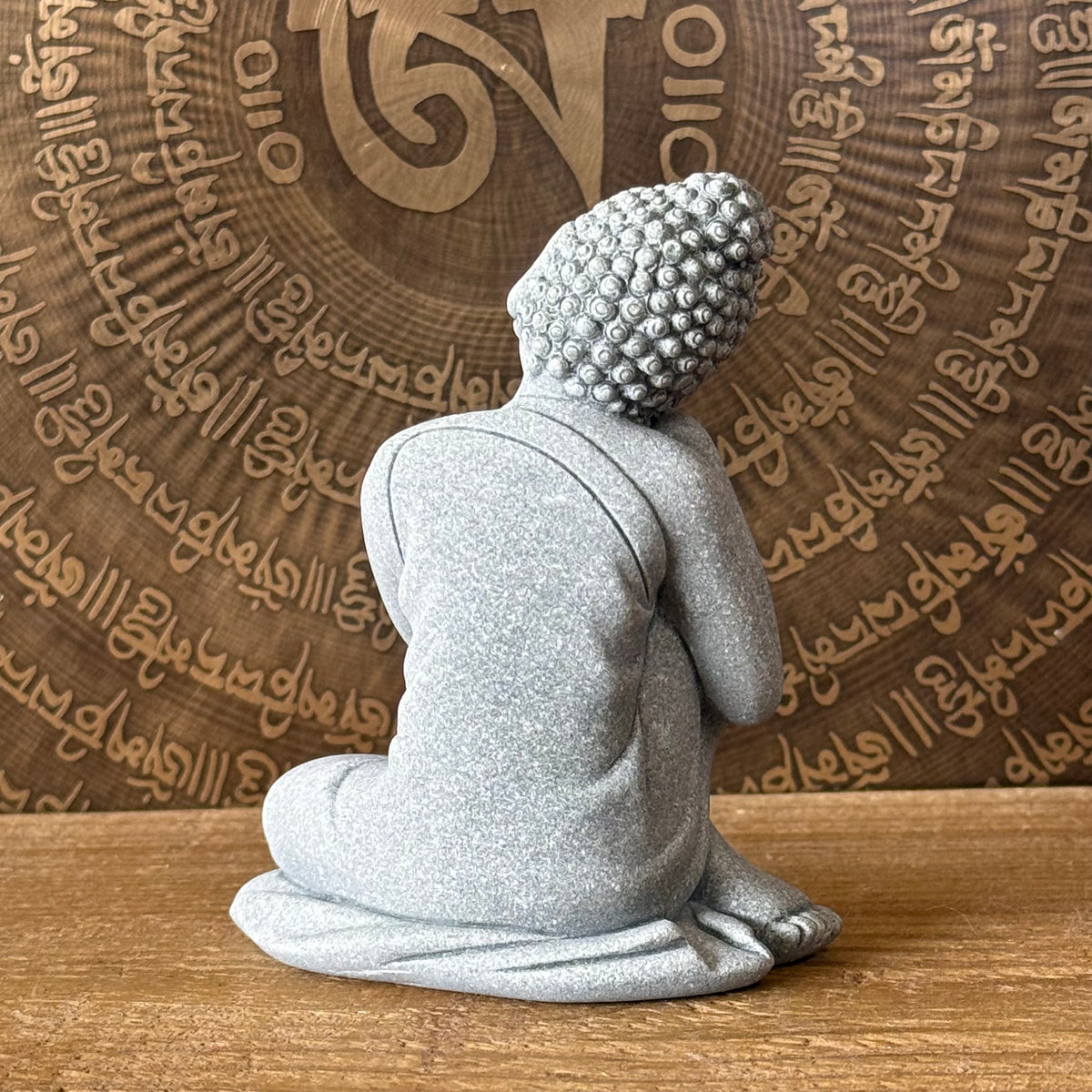 A gray glittery Buddha statue shown from the back, seated cross-legged on a wooden surface with a decorative wooden background featuring script and a central symbol.