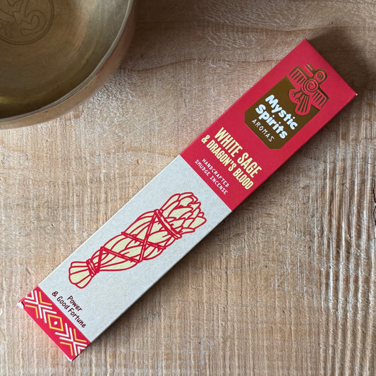 A rectangular product box for Mystic Spirits Artisan White Sage & Ashwagandha Steam Infused Oil, placed diagonally on a light wood grain surface next to a brass bowl.