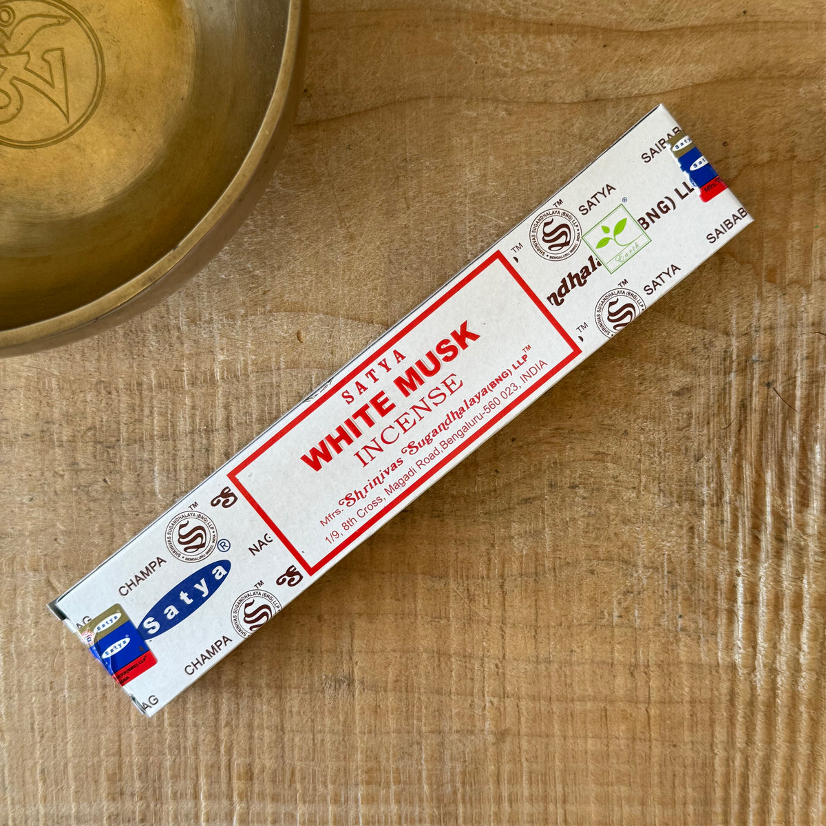 A single stick of Satya White Musk Incense placed diagonally on a light wood surface next to a brass bowl. The packaging is white with red and black text and logos.