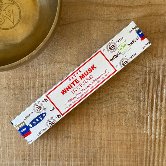 A single stick of Satya White Musk Incense placed diagonally on a light wood surface next to a brass bowl. The packaging is white with red and black text and logos.
