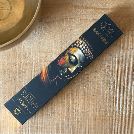 A long rectangular product box for BANJARA Buddha Wisdom incense sticks lies diagonally on a light wood surface. The box is dark navy blue with a printed Buddha face illustration in gold, orange, and white tones, plus gold text reading 'BANJARA' and 'BUDDHA Wisdom'.