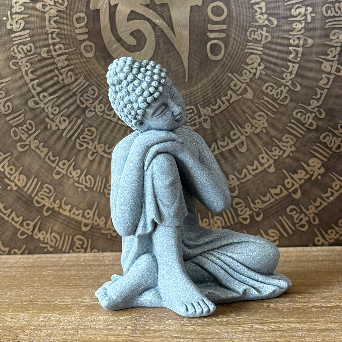 A small seated Buddha figurine in a light blue-gray stone-like finish, placed on a wooden surface in front of a circular brown backdrop with engraved script.