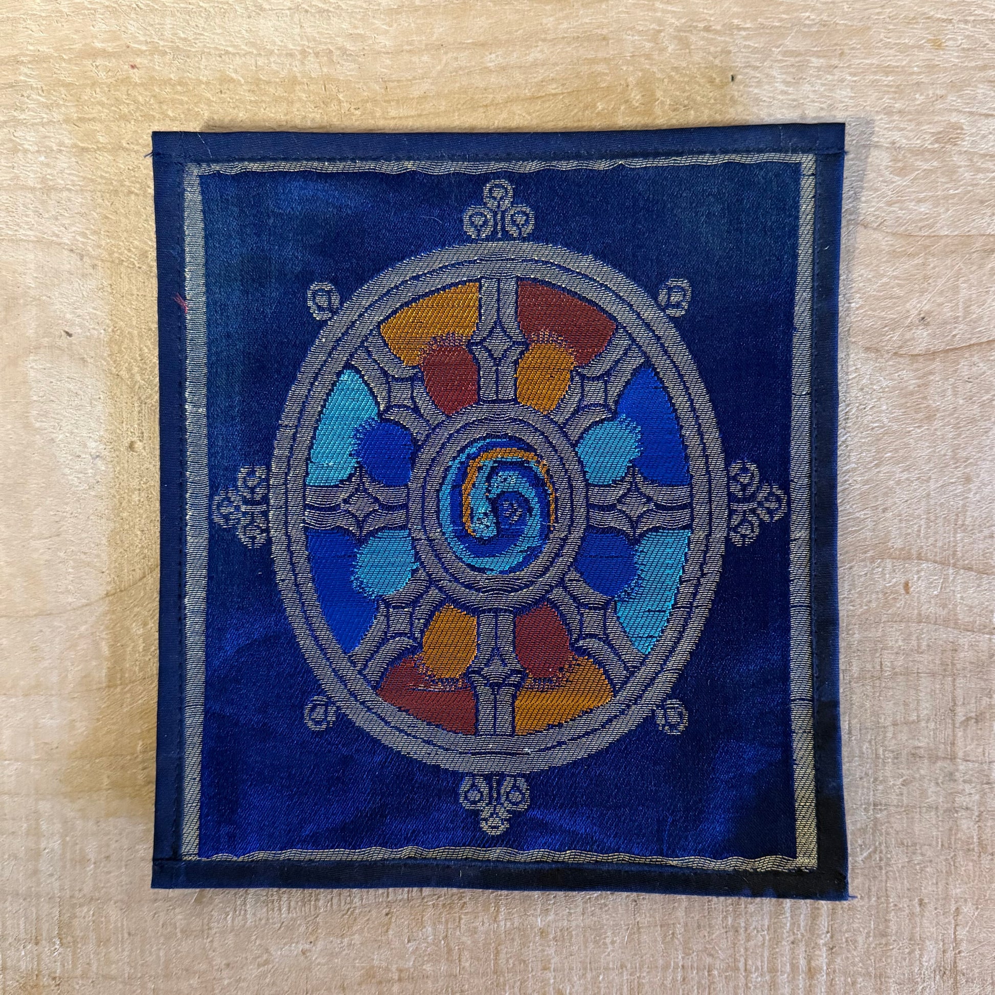 This Mini Tibetan altar cloth features a Dorje and a bold Dharma Wheel, symbolizing love, compassion, and spiritual protection. Handcrafted from thick silk brocade, this silk brocade altar mat is perfect for placement under a singing bowl, standing Dorje, or other sacred Buddhist ornaments.