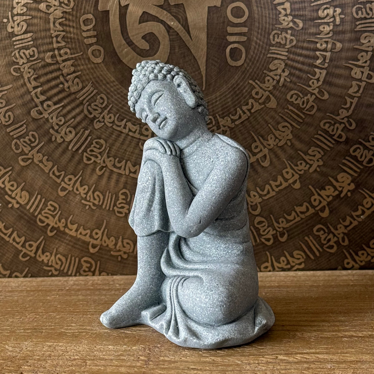 A gray stone Buddha statue in a kneeling pose with hands clasped under his arm, set on a wooden surface against a brown background with engraved script.