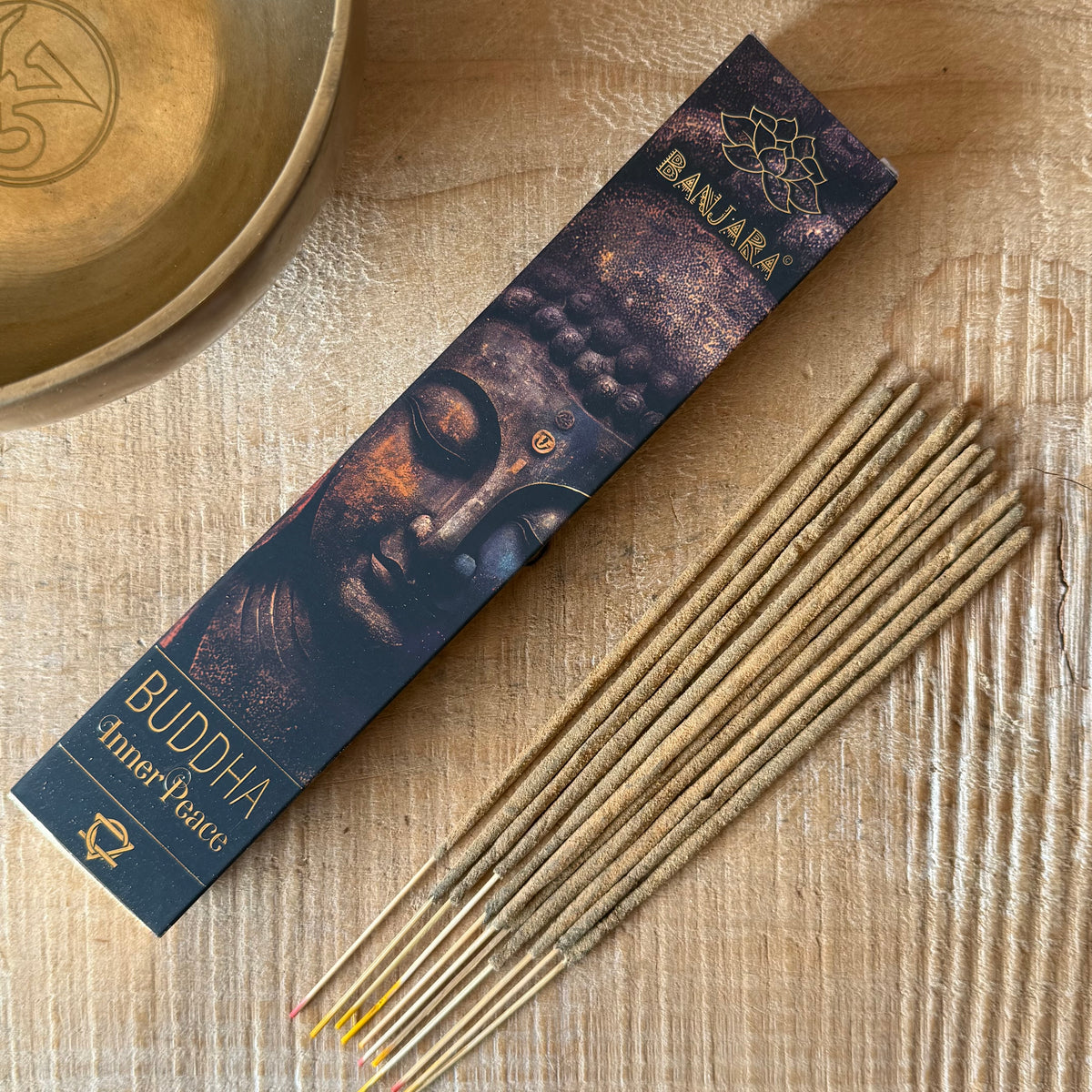 A box of Buddha Amher Peatus incense sticks lies diagonally on a light wood surface next to several unboxed incense sticks. The packaging features a dark navy blue background with a printed image of Buddha's face and gold text reading 'BAJRA' and 'BUDDHA Amher Peatus'.