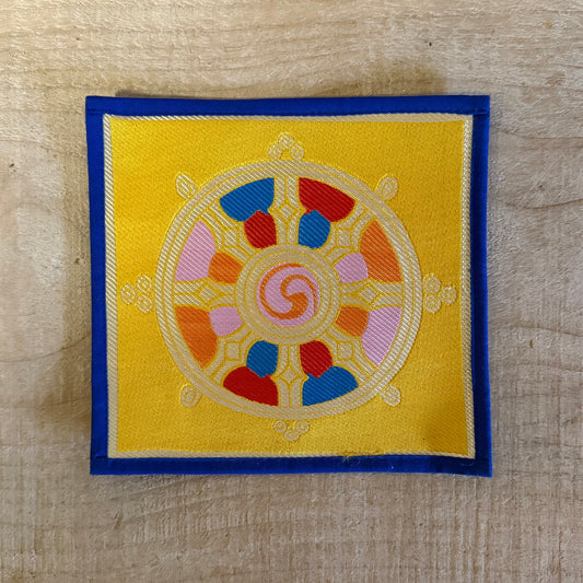 This Mini Tibetan altar cloth features a Dorje and a bold Dharma Wheel, symbols of love, compassion, and spiritual protection. Handcrafted from thick silk brocade, this silk brocade altar mat is ideal for placement under a singing bowl, standing Dorje, or other sacred Buddhist ornaments.