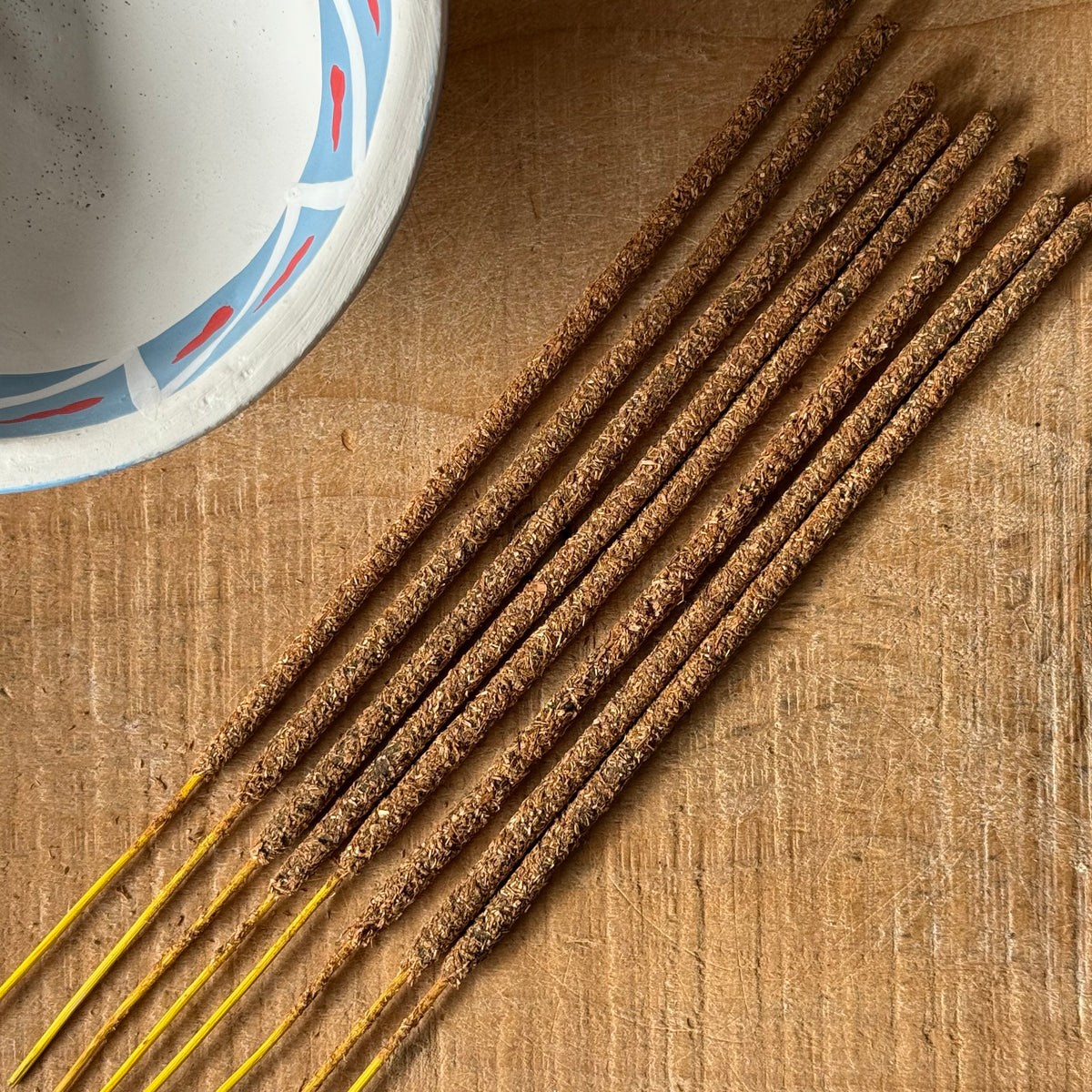 Several brown incense sticks with yellow tips are arranged diagonally on a wooden surface, with part of a white ceramic bowl visible in the top left corner.