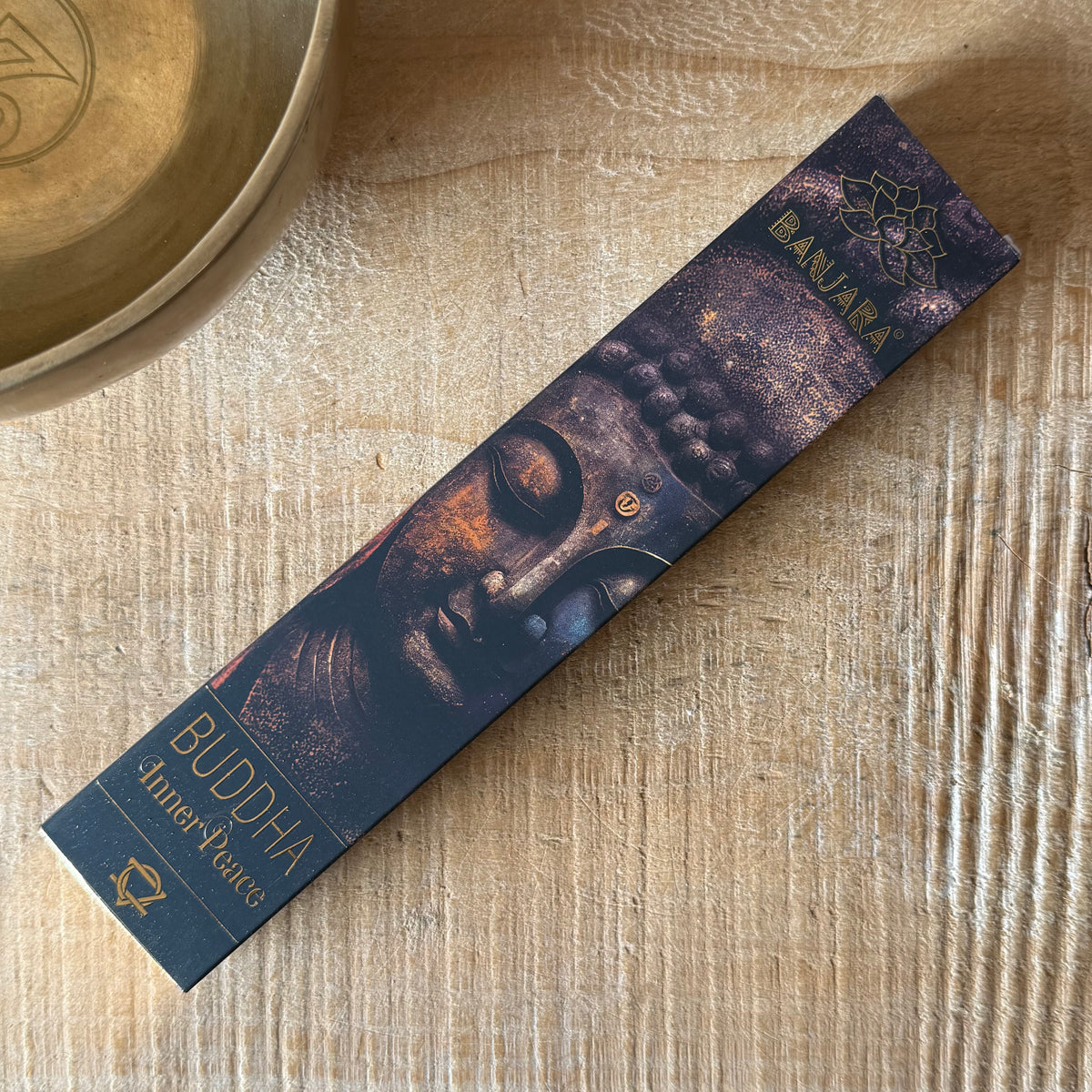 A rectangular incense stick box labeled 'Buddha AmperFume' and 'BAILARA' lies diagonally on a light wood surface next to a brass bowl.