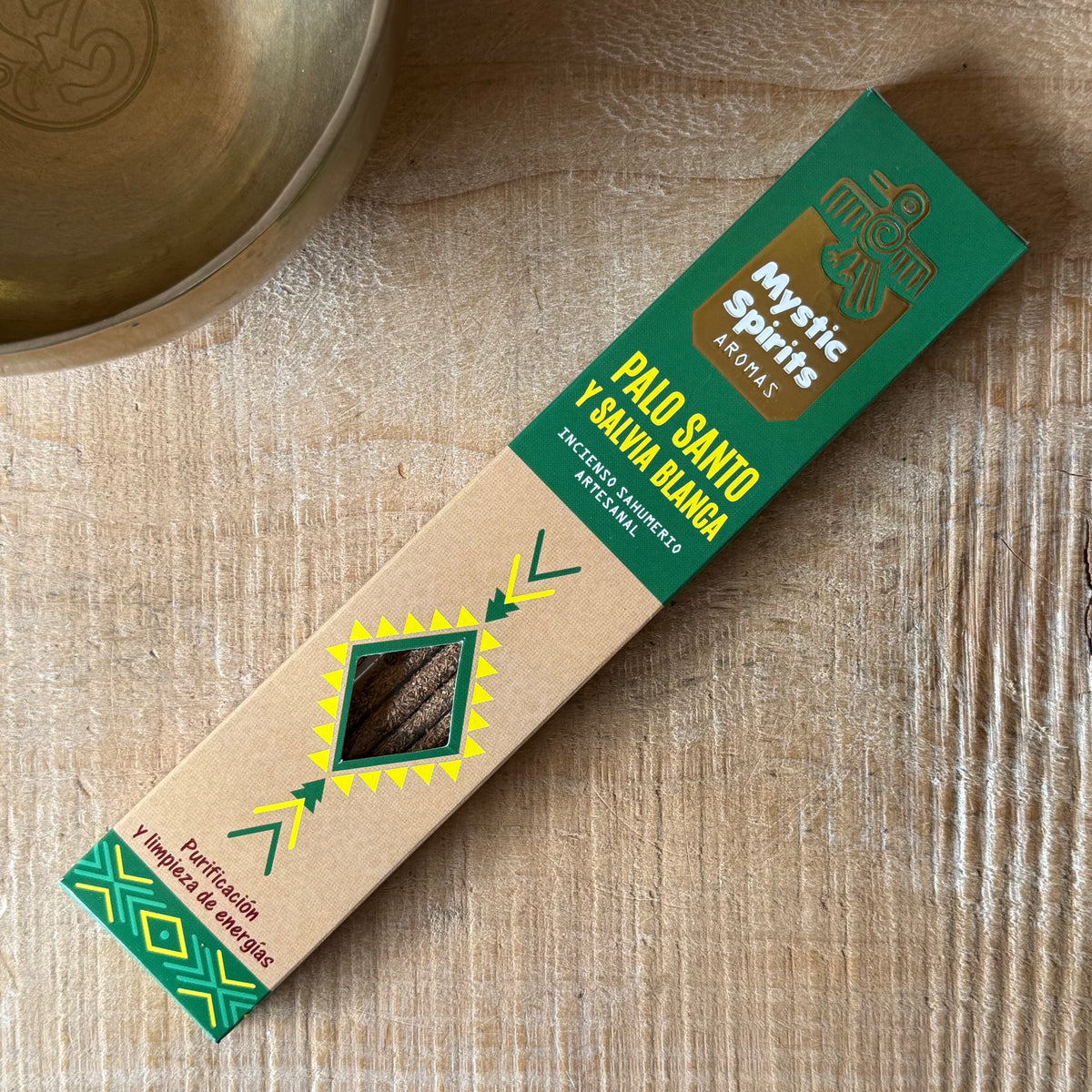 A package of Mystic Spires Palo Santo incense sticks on a light wood surface next to a brass bowl. The packaging is a combination of green and kraft paper with geometric designs.