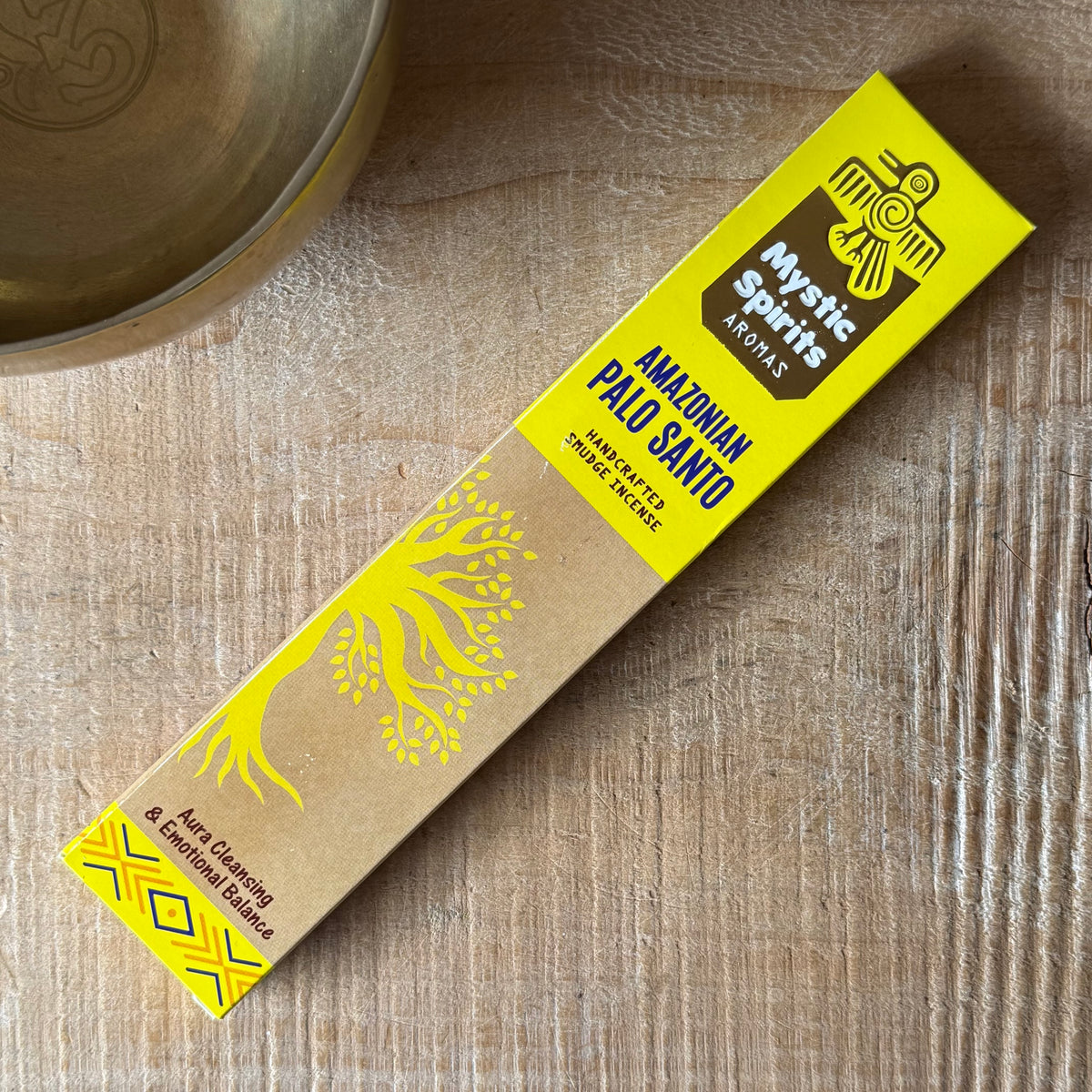 A box of Amazonian Palo Santo incense sticks by Mystic Spirits Aromas, placed diagonally on a light wood surface next to a brass bowl.
