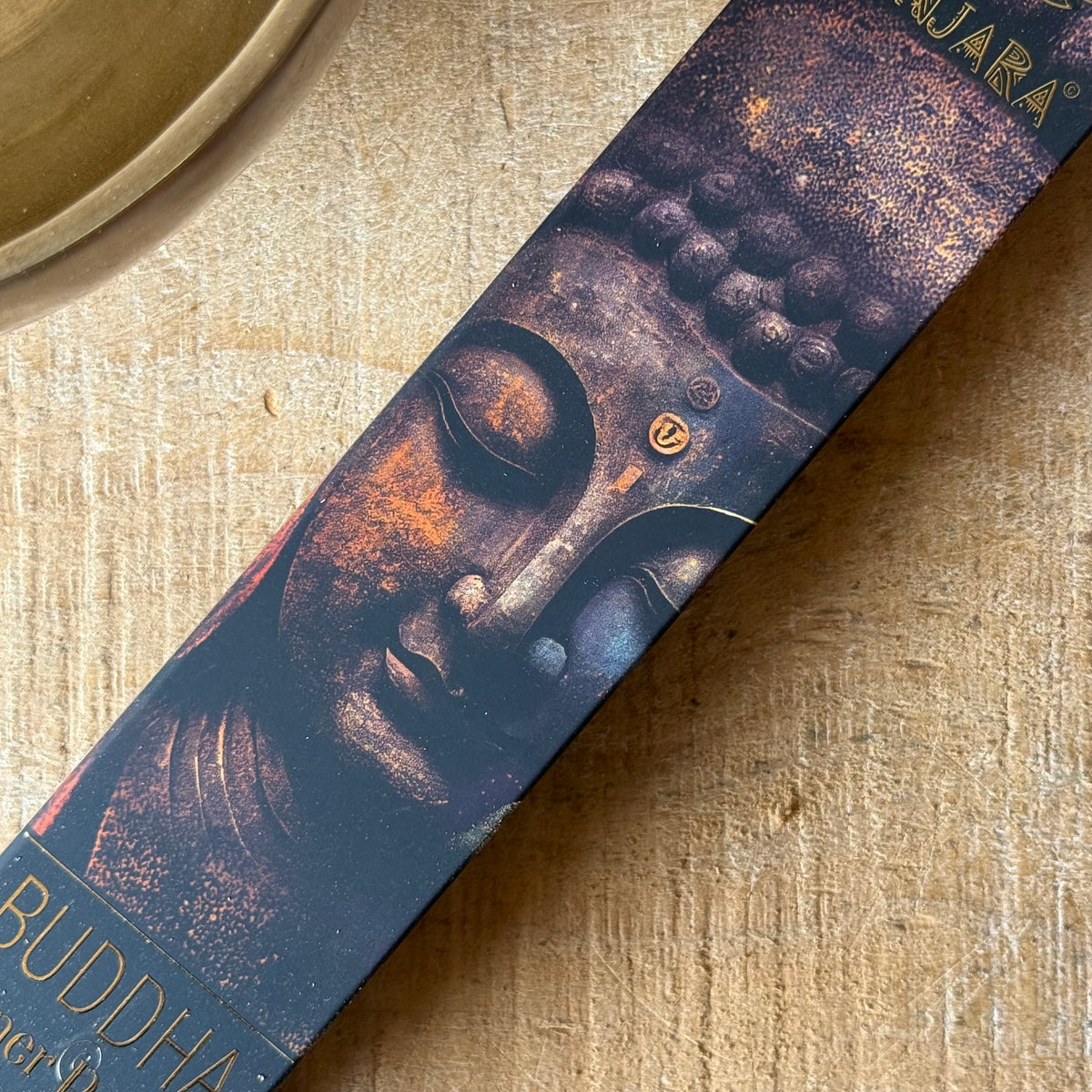 A close-up of a rectangular product box featuring a Buddha face illustration in dark blue, bronze, and gold tones, placed diagonally on a light wood surface.