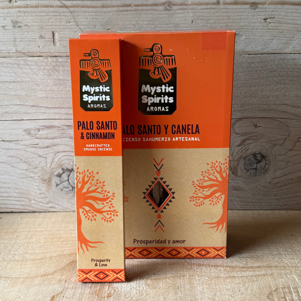Two boxes of Mystic Spirits Aromas Palo Santo incense sticks, one for 'Palo Santo & Cinnamon' and one for 'Palo Santo y Canela', displayed on a light wooden surface against a rustic wood plank background.