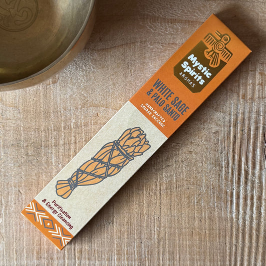 A box of Mystic Spirits Aromas White Sage & Palo Santo hand-cut sage and incense sticks, placed diagonally on a light wood tabletop next to a brass bowl.