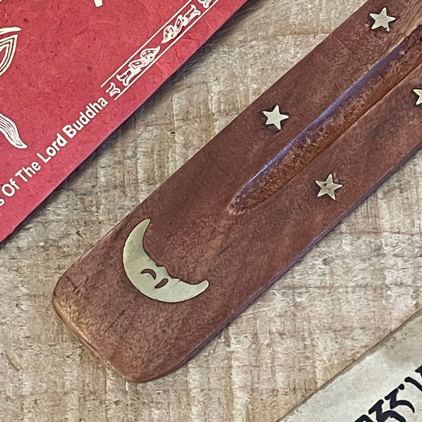 This wooden incense holder is a beautiful ash catcher with a celestial design, ideal for enhancing the atmosphere of any space. Crafted from natural wood and inlaid with a brass moon and stars pattern, this piece is both functional and decorative. Its slightly curved design holds stick incense securely while catching falling ash, keeping your surroundings clean.