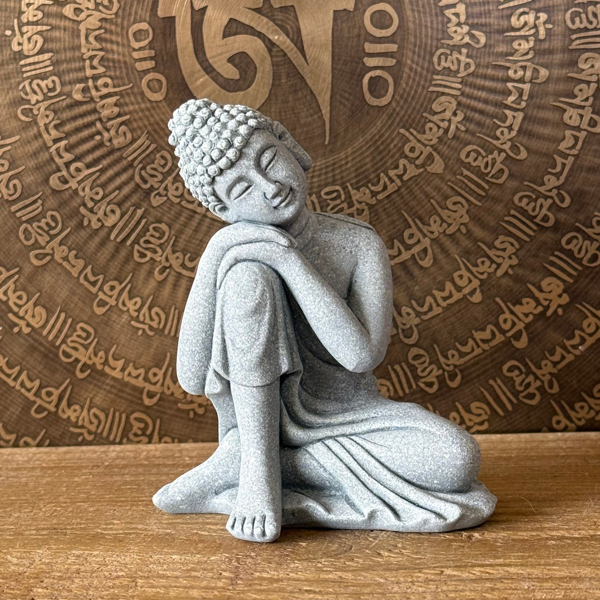 A gray stone Buddha statue in a seated meditation pose, placed on a wooden surface with a decorative bronze-toned circular backdrop featuring engraved script.