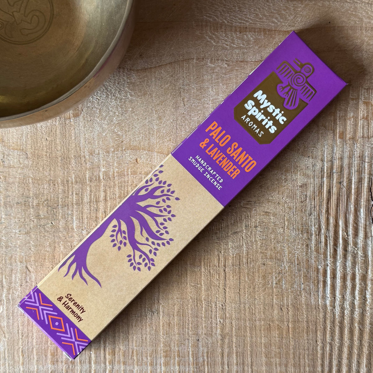 A rectangular box of Mystic Spirits Aromas Palo Santo & Lavender incense sticks lies diagonally on a light wood tabletop, with part of a brass bowl visible in the upper left corner.