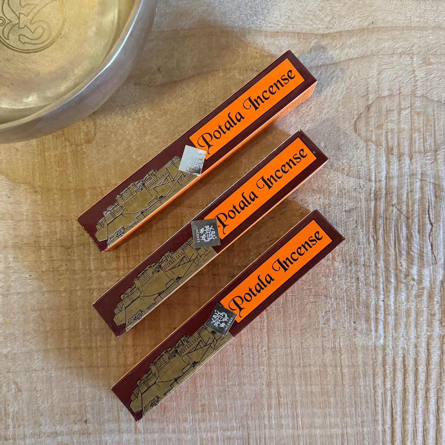 Three Potala Incense sticks on a wooden surface with a metal bowl in the background.