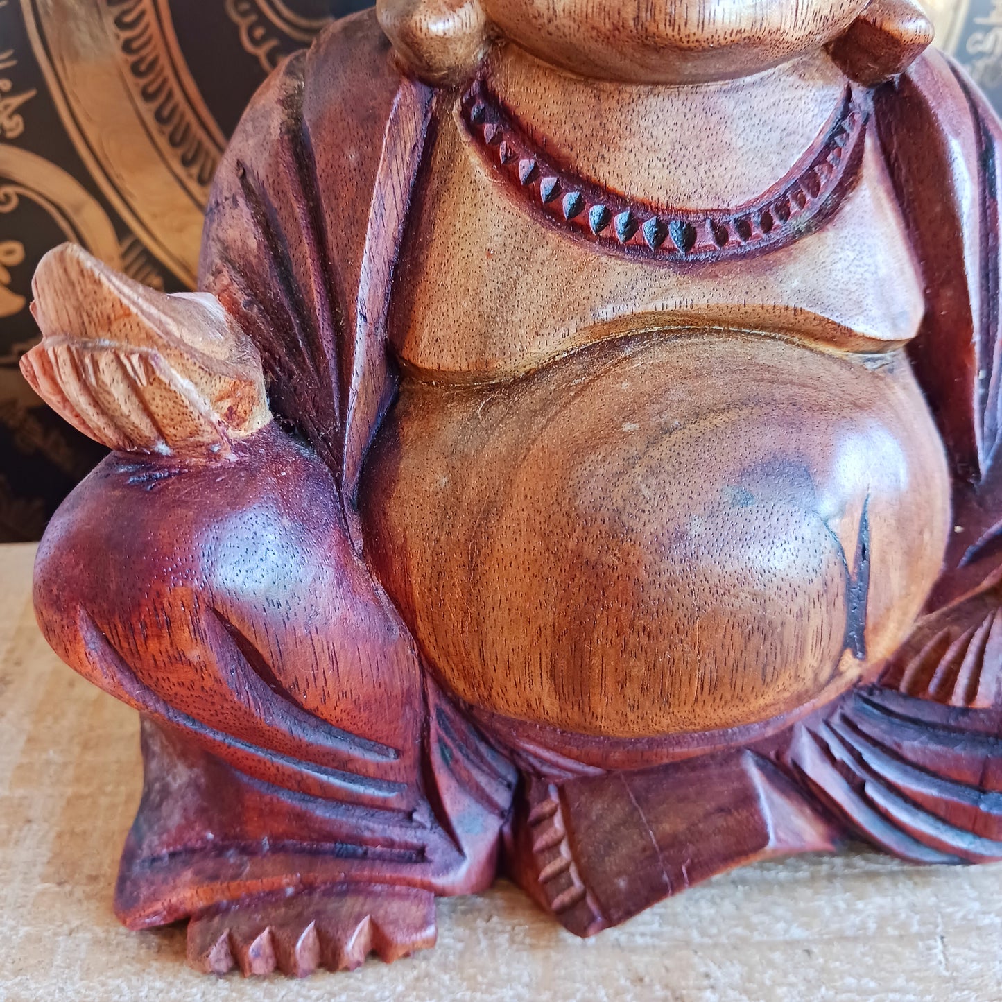 This charming Hand Carved Laughing Buddha is seated in a relaxed pose with a broad, joyful grin and rounded belly, beautifully carved from warm two-tone wood. The natural wood grain is enhanced with a protective varnish, highlighting the intricate details of his robes, prayer beads and expressive face.
