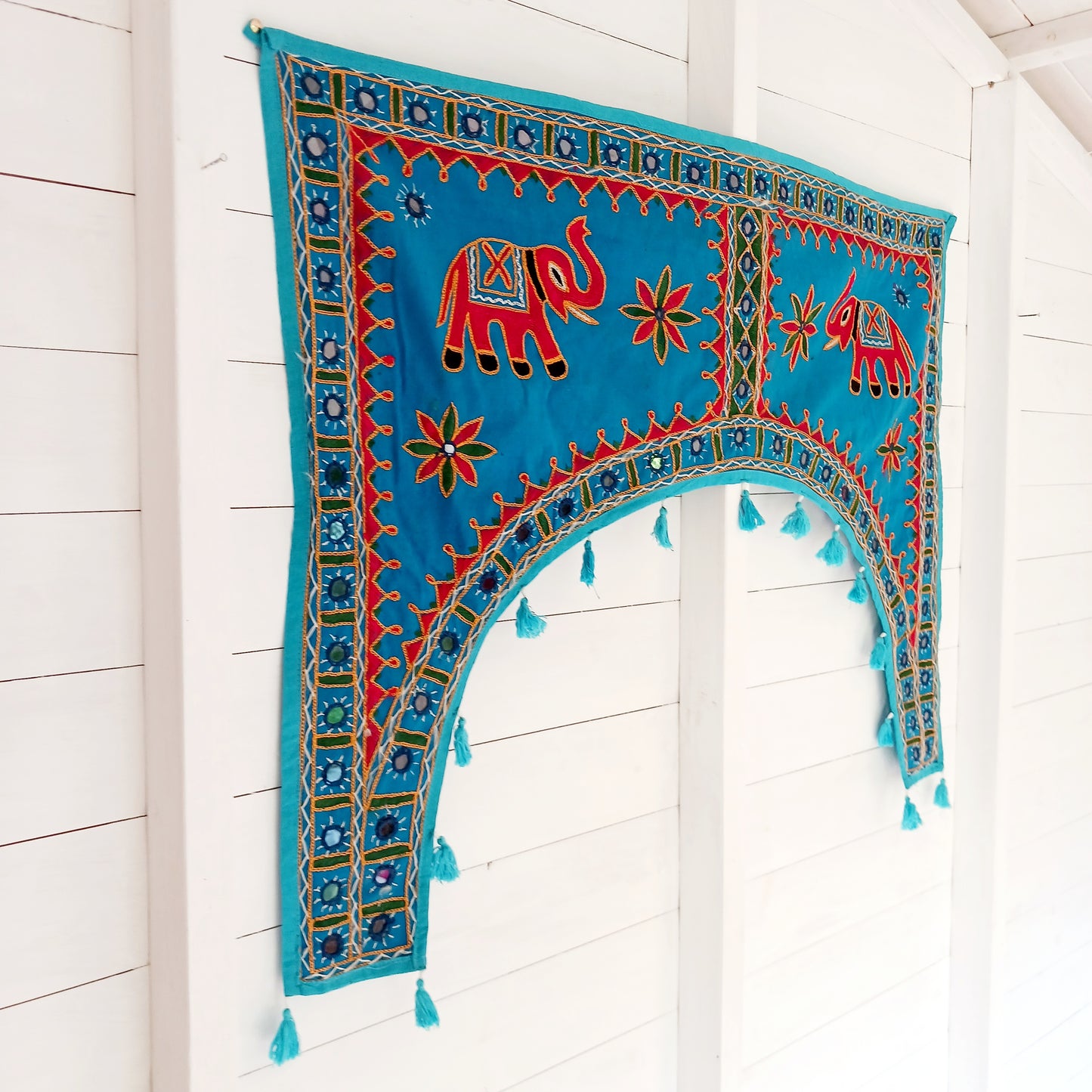 This beautiful turquoise Indian arch toran features two orange elephants with flowers either side and is adorned with mirrors and finished with tassels. This lovely toran has been meticulously hand-sewn and beautifully finished.  It really is a unique work of art.