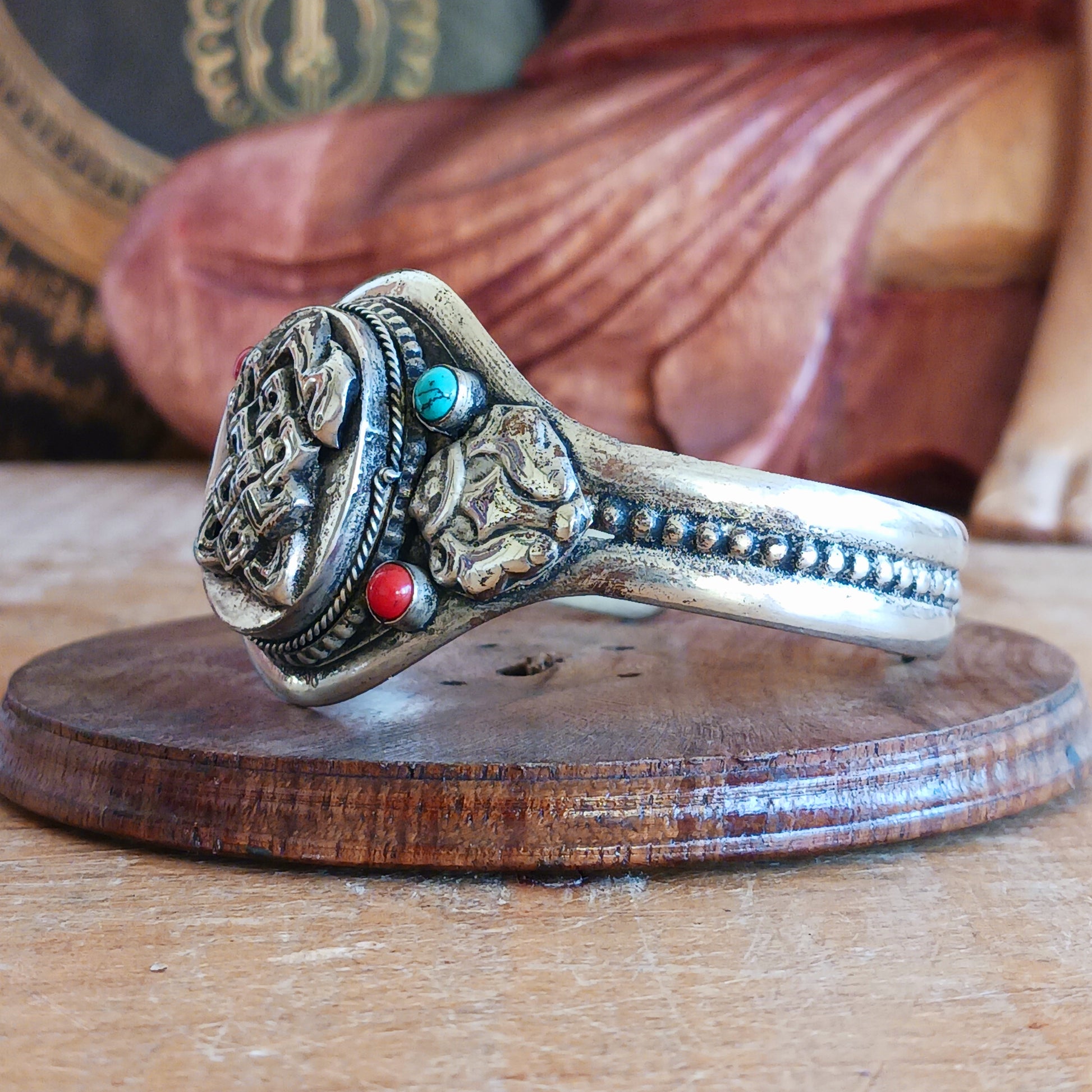 Embrace timeless elegance with this Endless Knot Tibetan Charm Bangle. Crafted from high-quality white metal and adorned with vibrant turquoise and coral semi-precious stones, this adjustable bangle showcases the sacred Endless Knot symbol on the front.