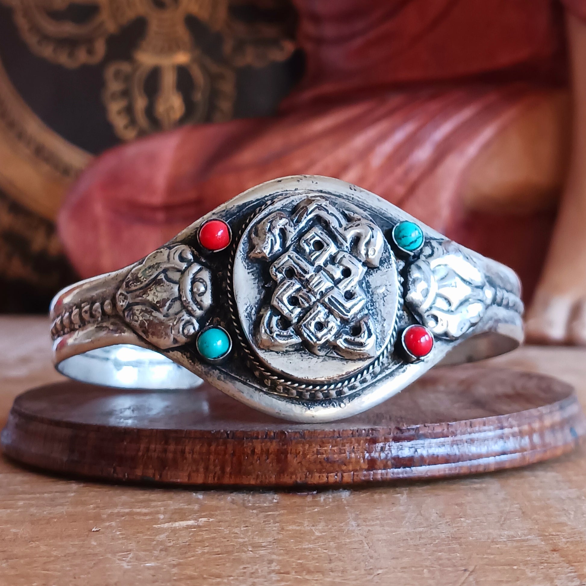 Embrace timeless elegance with this Endless Knot Tibetan Charm Bangle. Crafted from high-quality white metal and adorned with vibrant turquoise and coral semi-precious stones, this adjustable bangle showcases the sacred Endless Knot symbol on the front.