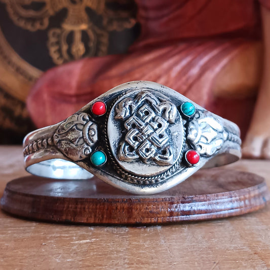 Embrace timeless elegance with this Endless Knot Tibetan Charm Bangle. Crafted from high-quality white metal and adorned with vibrant turquoise and coral semi-precious stones, this adjustable bangle showcases the sacred Endless Knot symbol on the front.