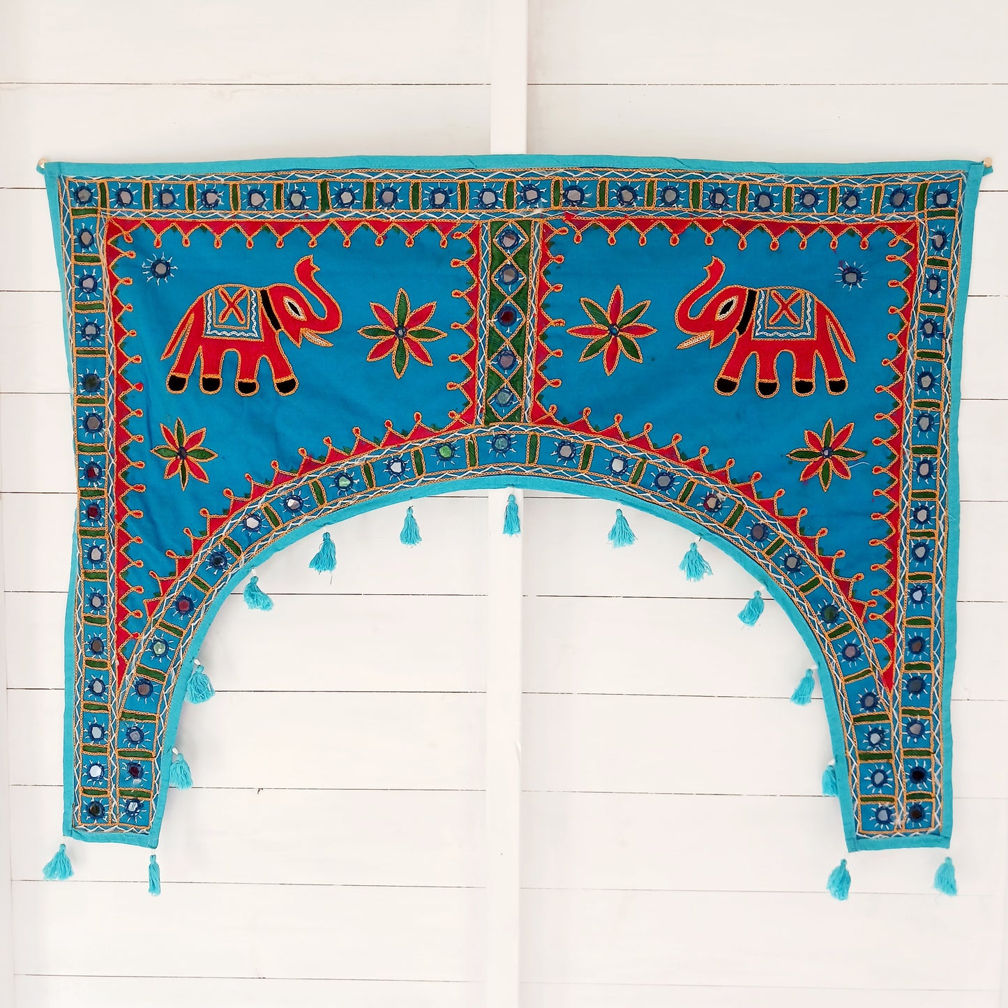 This beautiful turquoise Indian arch toran features two orange elephants with flowers either side and is adorned with mirrors and finished with tassels. This lovely toran has been meticulously hand-sewn and beautifully finished.  It really is a unique work of art.