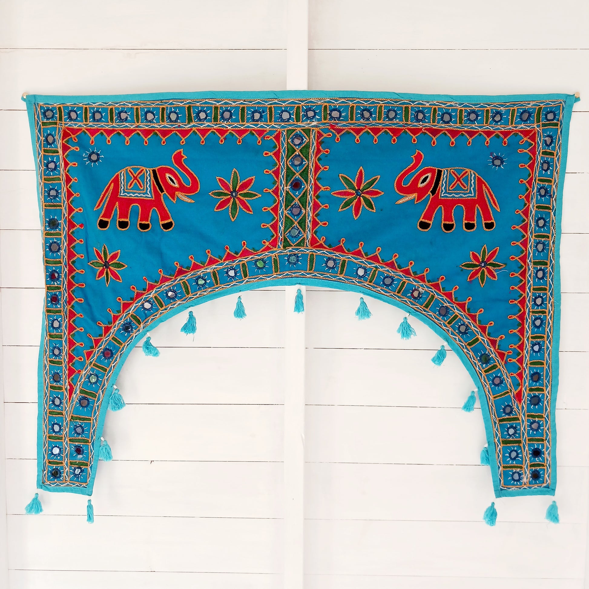 This beautiful turquoise Indian arch toran features two orange elephants with flowers either side and is adorned with mirrors and finished with tassels. This lovely toran has been meticulously hand-sewn and beautifully finished.  It really is a unique work of art.