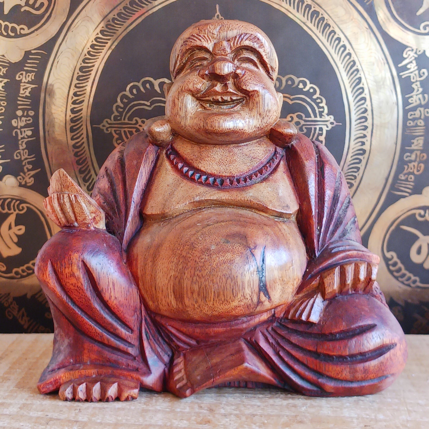 This charming Hand Carved Laughing Buddha is seated in a relaxed pose with a broad, joyful grin and rounded belly, beautifully carved from warm two-tone wood. The natural wood grain is enhanced with a protective varnish, highlighting the intricate details of his robes, prayer beads and expressive face.