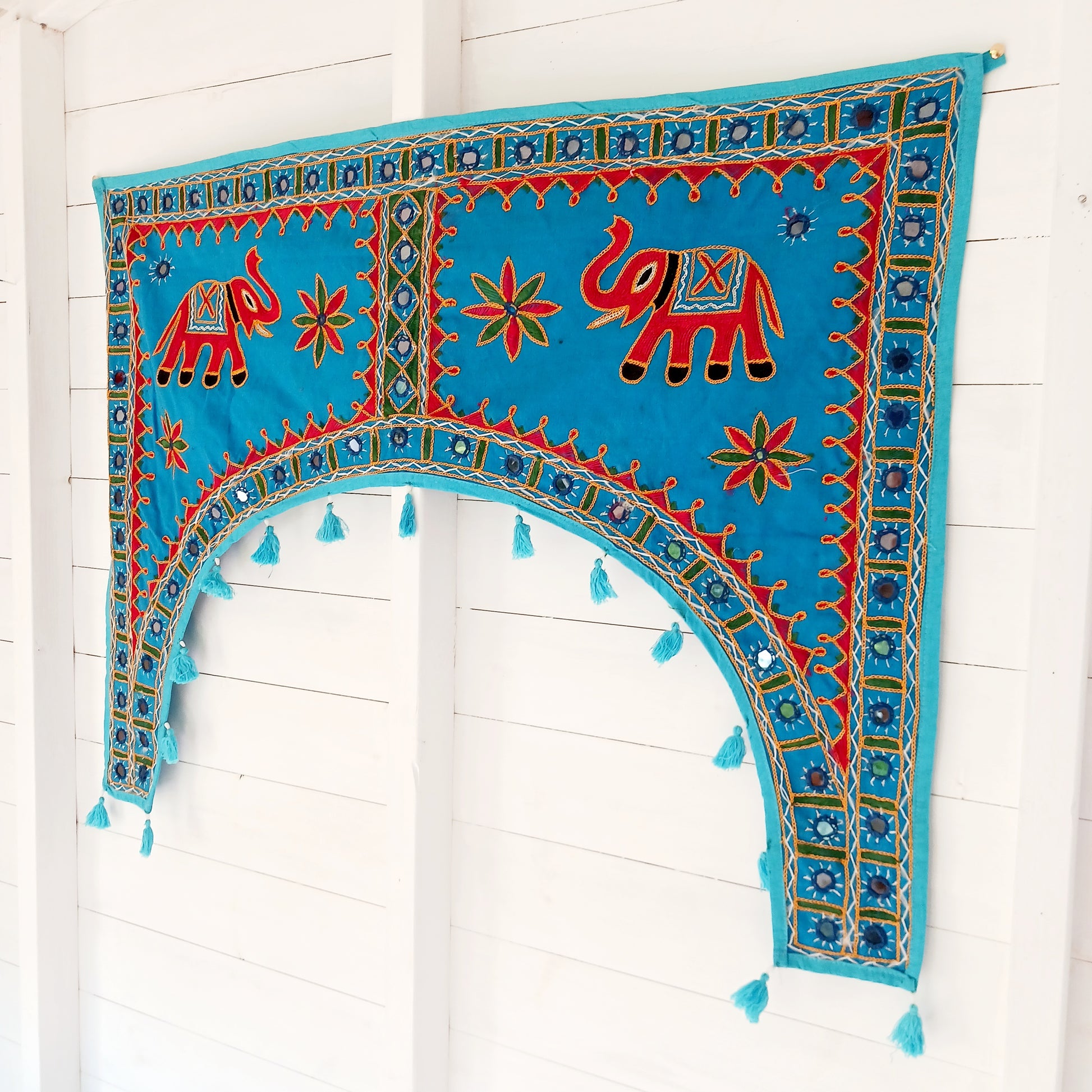 This beautiful turquoise Indian arch toran features two orange elephants with flowers either side and is adorned with mirrors and finished with tassels. This lovely toran has been meticulously hand-sewn and beautifully finished.  It really is a unique work of art.